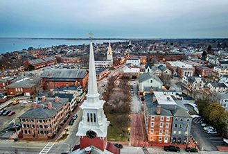Aerial view of a small coastal town with a prominent white church steeple in the foreground, surrounded by historic brick buildings and a calm harbor.