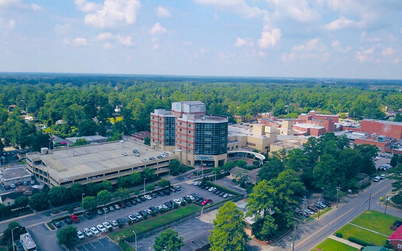 Aerial view of a hospital complex surrounded by trees. The main building features a distinct cylindrical glass section. Cars are parked below, under a clear blue sky.