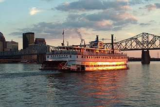 A white steamboat with glowing lights sails on a river at sunset. A city skyline and a large, arched bridge are seen in the background.