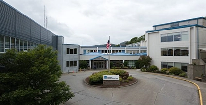 Peace Health Ketchikan location, A large, modern hospital building with a U.S. flag on the roof. The entrance is surrounded by greenery and a sign with the hospital's name. Overcast sky above.