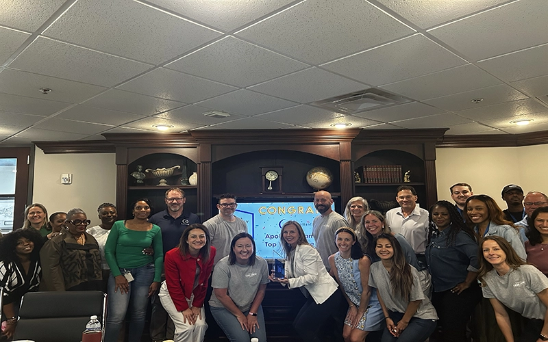 A diverse group of people smiles and gathers in an office, celebrating in front of a congratulatory screen. The atmosphere is joyful and collaborative.