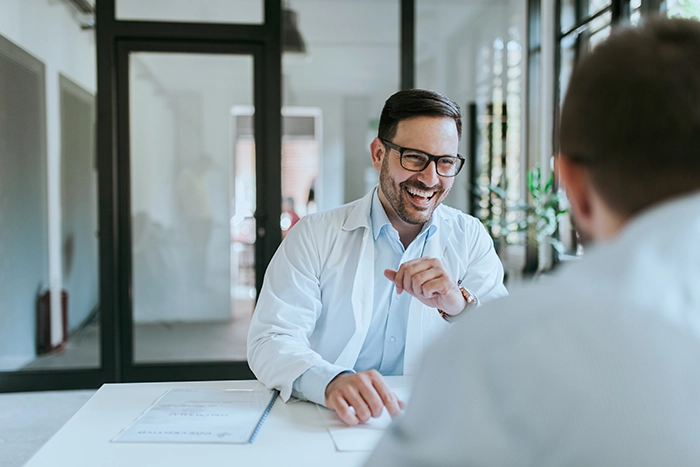 A smiling doctor in a white coat, with a stethoscope, sits at a desk facing a patient. The setting conveys a friendly, professional atmosphere.