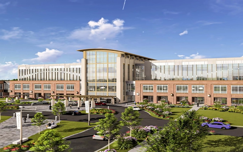 Modern hospital building with large glass windows, surrounded by landscaped gardens and a parking area under a bright blue sky with fluffy clouds.