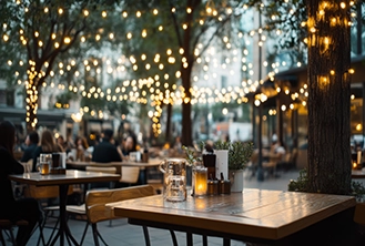 Outdoor cafe with wooden tables under string lights, surrounded by trees. People dine in the background, creating a cozy, warm atmosphere.