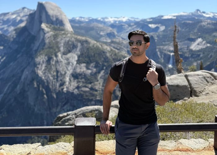 Dr.Raj, in sunglasses stands by a railing with a mountain backdrop, exuding a relaxed and adventurous vibe. He's wearing a black t-shirt and backpack.