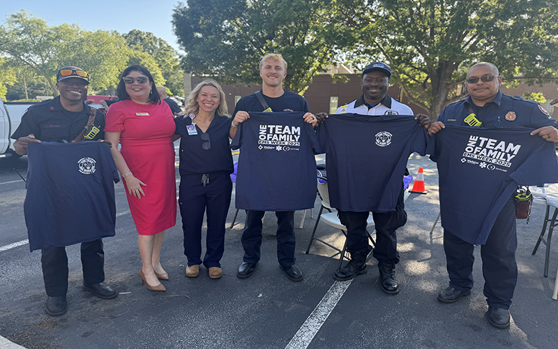 A group of six smiling people stand outdoors, holding matching navy 