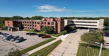 Aerial view of a brick office building and parking lot, surrounded by trees, under a clear blue sky. The scene conveys a calm, professional atmosphere.