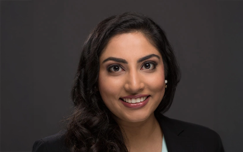 Amna Jamshad, Smiling woman with long, wavy dark hair, wearing a black blazer and light top, against a dark background. She conveys confidence and warmth.