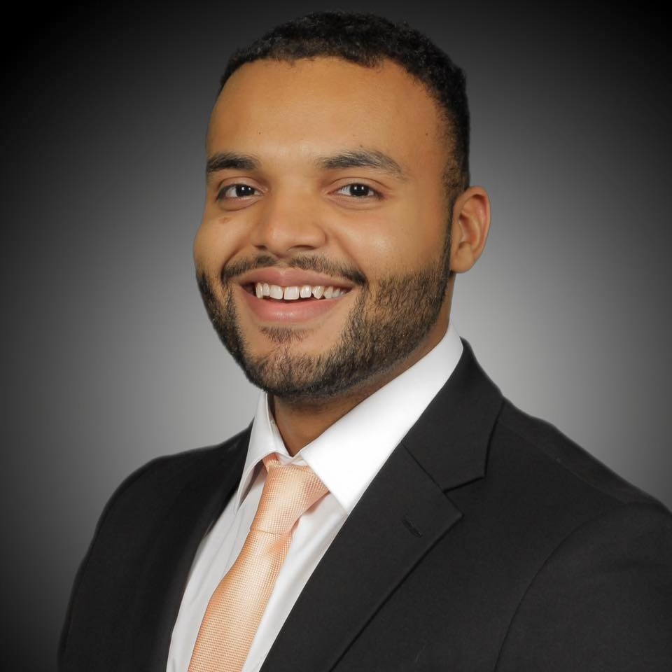A warm, professional portrait of professional. He has short hair, is wearing a blazer and tie, and is smiling directly at the camera against a neutral backdrop