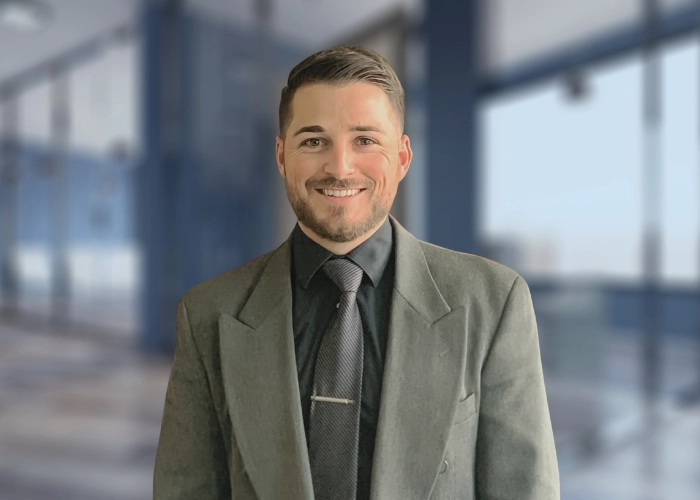 A smiling man in a gray suit and black shirt stands in a modern office hallway. The setting is bright and professional, exuding a confident and welcoming tone.