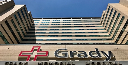 Upward view of Grady Memorial Hospital's facade with beige color and dark windows. Prominent red cross logo and 