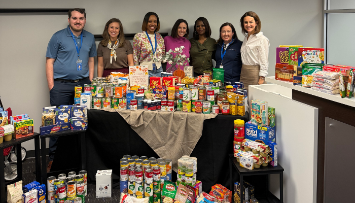 A group of seven people stand smiling behind a table filled with canned goods and packaged foods for a food drive. The atmosphere is cheerful and supportive.