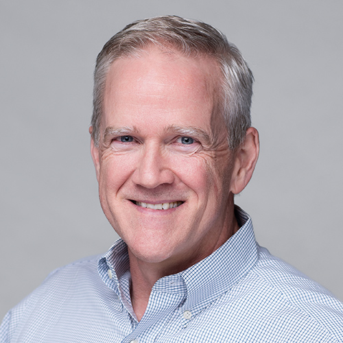 A smiling man with short gray hair wears a light blue checkered shirt. The background is a neutral gray, conveying a professional and approachable tone.