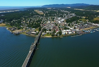 Aerial view of a bridge crossing a wide river, leading to a small town with clustered buildings and surrounded by dense green forests and hills.