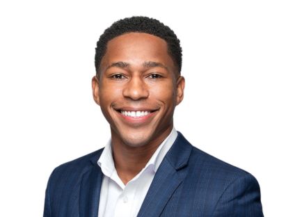 Professional, Dany Accilien in a blue suit and white shirt smiles warmly at the camera. He has short, curly hair and stands against a plain white background