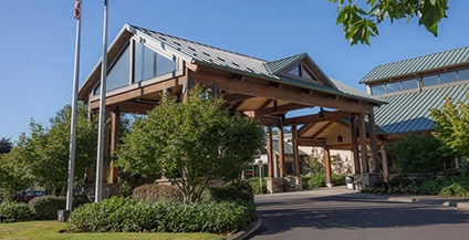 Modern building of Peace Health at Cottagegrove location with a green, gabled roof, large wooden beams, and glass panels, surrounded by lush greenery under a clear blue sky. Calm atmosphere.
