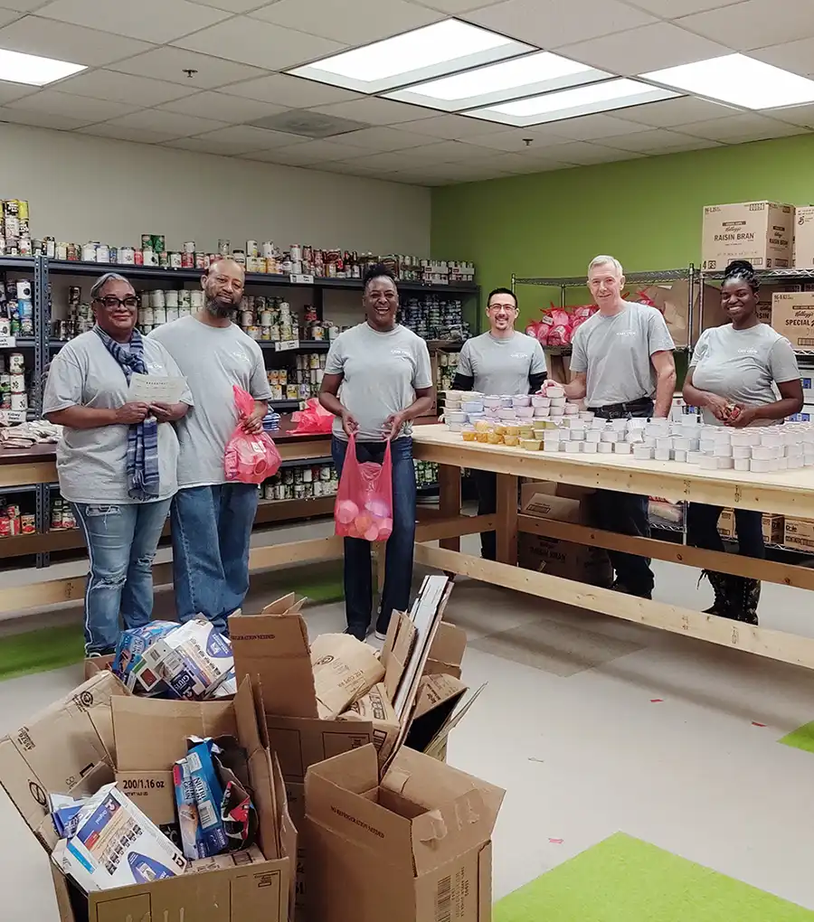 A group of six smiling individuals in gray shirts volunteering at a food bank, organizing canned goods and packing items in red bags. Shelves are stocked behind them.