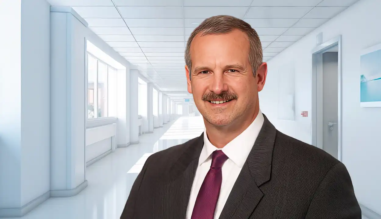 A man smiling at the camera, he has on a dark purple tie and gray suit, the background is an empty medical hallway